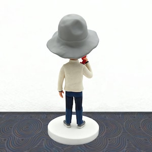 May include: A figurine of a person wearing a light gray hat, a cream-colored sweater, dark blue jeans, and gray shoes. The figure is holding a red object. The figurine is standing on a white circular base.