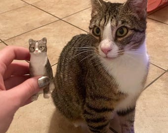 May include: A brown tabby cat with white markings sits on a tiled floor, looking at a small figurine of a cat in a person's hand.