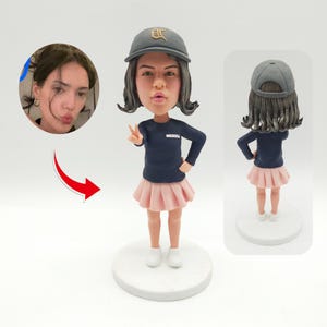 Op de afbeelding: Een gepersonaliseerd bobblehead-beeldje van een persoon die een zwart-witte baseballpet, een donkerblauw shirt met lange mouwen, een roze rok en witte sneakers draagt. Het beeldje staat op een witte voet.