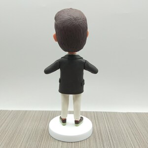 May include: A figurine of a person viewed from the back, wearing a black jacket, white pants, and brown shoes with green soles. The figure has brown hair and is standing on a white circular base.