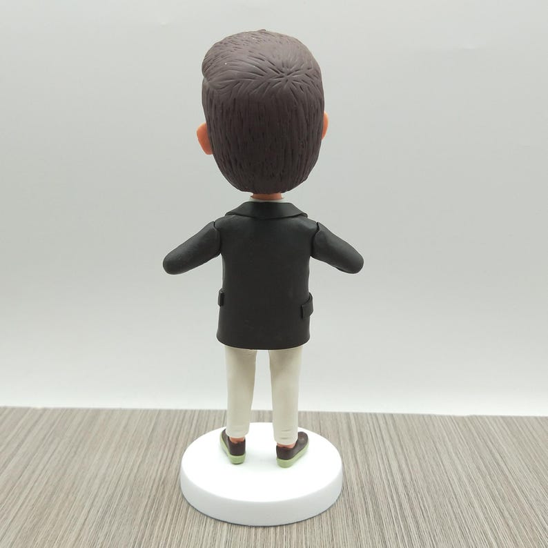 May include: A figurine of a person viewed from the back, wearing a black jacket, white pants, and brown shoes with green soles. The figure has brown hair and is standing on a white circular base.