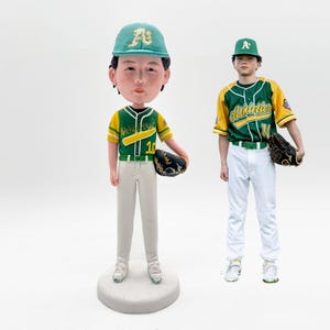 Könnte beinhalten: Eine Bobblehead-Figur eines Baseballspielers in grün-gelber Uniform und Kappe, die einen Baseballhandschuh hält. Die Figur steht auf einem runden, grauen Sockel. Eine Person in ähnlicher Uniform und Kappe steht daneben.