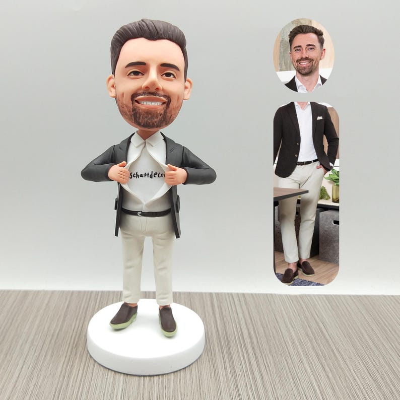May include: A custom bobblehead figurine of a man in a suit, pulling open his shirt to reveal the text "Schatttdeco". The figure is on a white base, wearing a black blazer, white shirt, and beige pants. A smaller image of the man is in the background.