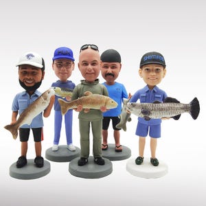 May include: A collection of six bobblehead figurines of men holding fish. Each figure is dressed in casual attire, wearing hats, and standing on a circular base. The fish vary in size and color, with details like scales and fins.
