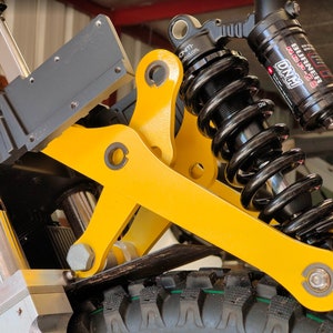 May include: Close-up of a yellow motorcycle suspension system with a black coil spring and a black shock absorber. The shock absorber has the text "BURNER" and "DNM" printed on it.