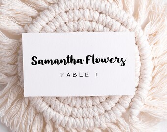 May include: White card with black text that reads "Samantha Flowers Table I" on a white macrame coaster with fringe.