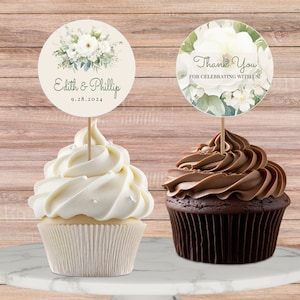 Könnte beinhalten: Zwei Cupcakes mit floralen Toppern. Ein Cupcake ist weiß mit weißem Frosting und der andere ist Schokolade mit Schokoladenfrosting. Der Topper auf dem weißen Cupcake sagt "Edith & Phillip 9.28.2024". Der Topper auf dem Schokoladencupcake sagt "Vielen Dank fürs Feiern mit uns!"