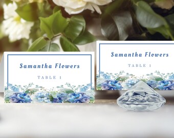 Può includere: Due segnaposto bianchi con bordi blu e disegni floreali. Le carte riportano "Samantha Flowers" e "Table 1". Un oggetto trasparente a forma di diamante sfaccettato si trova di fronte alle carte. Lo sfondo comprende fiori e verde.
