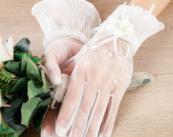 Puede incluir: Guantes de novia blancos con ribete de encaje y adornos de perlas. Los guantes tienen un puño con volantes y un detalle de lazo.