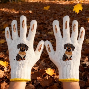 May include: A pair of white knit gloves with a dachshund dog graphic. The gloves have a yellow cuff and brown wheat stalk designs. The dog is wearing a sweater, and the name "John" is printed below the dog.