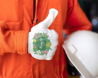 Op de afbeelding: Een witte werkhandschoen met een bloemenpatroon en de naam "Nancy" in het groen. De handschoen wordt gedragen door iemand in een oranje overall.