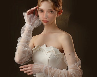 May include: White bridal gloves with pearl embellishments. The gloves are made of sheer tulle and have a ruffled cuff. The gloves are worn by a person with their arms extended.