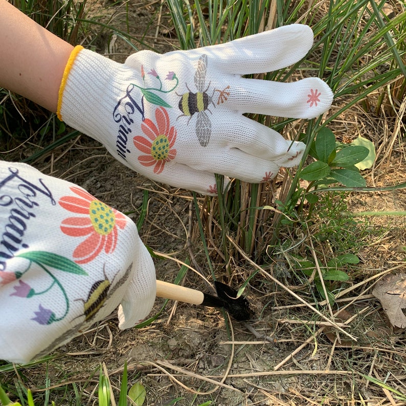 May include: A pair of white gardening gloves with a floral and bee design. The gloves have the text "Love, Grow" printed on them. The gloves are being used to weed a garden.
