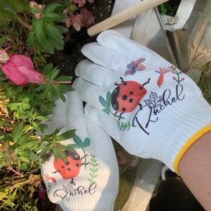 K&ouml;nnte beinhalten: Ein Paar wei&szlig;e Gartenhandschuhe mit gelbem Saum. Die Handschuhe haben ein Marienk&auml;fer- und Blumendesign und den Namen "Rachel" aufgedruckt.