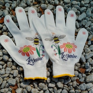May include: A pair of white gardening gloves with yellow trim. The gloves have a floral design with bees and the name "Lorraine" printed on each glove.