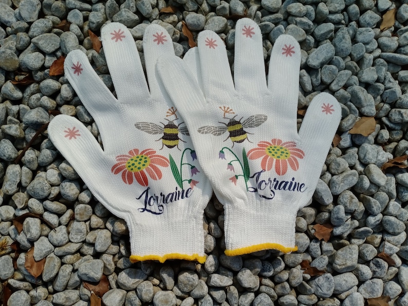 May include: A pair of white gardening gloves with yellow trim. The gloves have a floral design with bees and the name "Lorraine" printed on each glove.