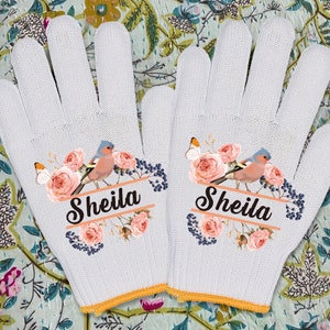Puede incluir: Un par de guantes de jardinería blancos con un diseño floral y el nombre "Sheila" impreso en cada guante. Los guantes presentan un pájaro rosa y azul con una mariposa y detalles florales.