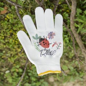 K&ouml;nnte beinhalten: Wei&szlig;er Gartenhandschuh mit einem Marienk&auml;fer- und Blumendesign. Der Handschuh tr&auml;gt den Namen "Rachel" aufgedruckt.