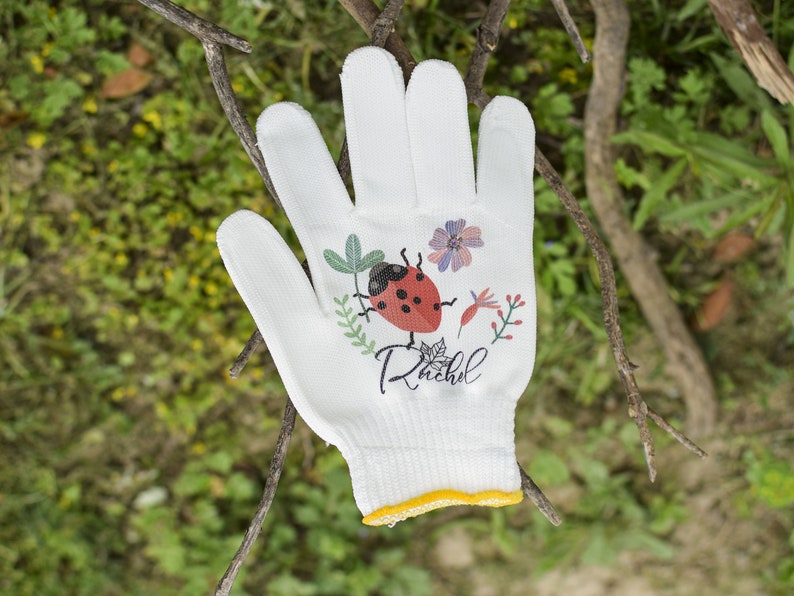 K&ouml;nnte beinhalten: Wei&szlig;er Gartenhandschuh mit einem Marienk&auml;fer- und Blumendesign. Der Handschuh tr&auml;gt den Namen "Rachel" aufgedruckt.