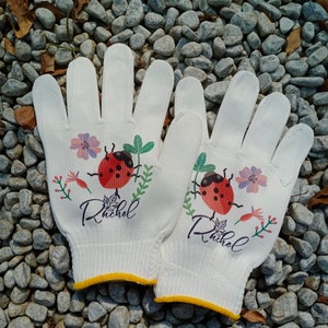 K&ouml;nnte beinhalten: Ein Paar wei&szlig;e Gartenhandschuhe mit einem Marienk&auml;fer- und Blumendesign. Die Handschuhe haben einen gelben Saum und der Name "Rachel" ist auf jedem Handschuh aufgedruckt.