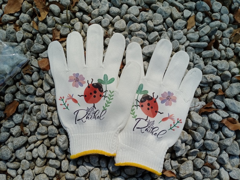K&ouml;nnte beinhalten: Ein Paar wei&szlig;e Gartenhandschuhe mit einem Marienk&auml;fer- und Blumendesign. Die Handschuhe haben einen gelben Saum und der Name "Rachel" ist auf jedem Handschuh aufgedruckt.