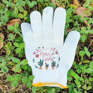 Op de afbeelding: Witte tuinhandschoenen met de tekst "Happy Mother's Garden" erop gedrukt. De handschoenen hebben een gele rand rond de pols.