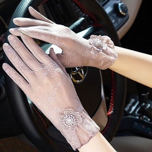 May include: A pair of delicate, sheer lace gloves with floral embellishments. The gloves are a light beige color and have a subtle pattern. The gloves are shown being worn by a person driving a car.