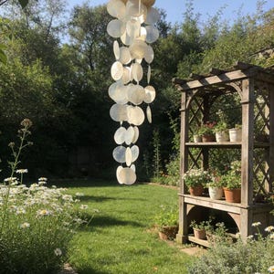 May include: A hanging wind chime with a wooden top and numerous, round, iridescent shell discs. The chime hangs in a garden setting with green grass, flowers, and a wooden trellis.