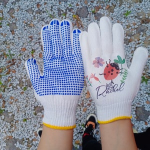 K&ouml;nnte beinhalten: Zwei Paar wei&szlig;e Baumwoll-Arbeitshandschuhe. Ein Paar hat einen blauen Punktgriff und das andere hat ein Marienk&auml;fer- und Blumendesign mit dem Namen Rachel darauf gedruckt.