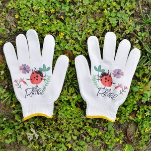 K&ouml;nnte beinhalten: Ein Paar wei&szlig;e Gartenhandschuhe mit einem Marienk&auml;fer- und Blumendesign. Die Handschuhe haben den Namen "Rachel" aufgedruckt.