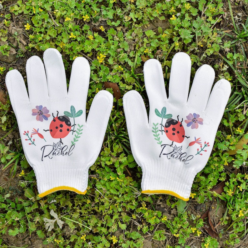 K&ouml;nnte beinhalten: Ein Paar wei&szlig;e Gartenhandschuhe mit einem Marienk&auml;fer- und Blumendesign. Die Handschuhe haben den Namen "Rachel" aufgedruckt.