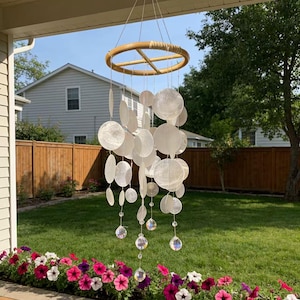 May include: A decorative wind chime featuring a bamboo ring and numerous white, circular shell discs. Clear crystal beads hang beneath the shells. The chime is suspended from a porch, with a garden and flowers in the background.