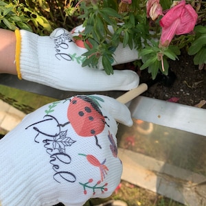 K&ouml;nnte beinhalten: Ein Paar wei&szlig;e Gartenhandschuhe mit einem Marienk&auml;fer- und Blumenmuster. Die Handschuhe haben den Schriftzug "Rachel" aufgedruckt.