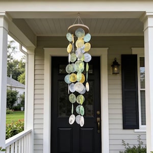 May include: A hanging wind chime with a wooden top and strands of iridescent, circular discs in shades of yellow, green, and blue. The wind chime hangs in front of a black door on a porch with white trim and a gray exterior.