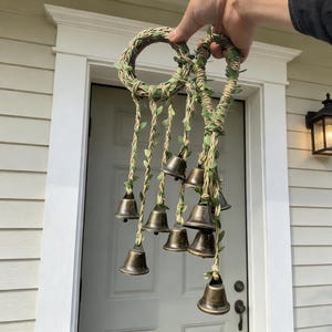 May include: A decorative wind chime with a woven ring and hanging bronze bells. The ring and hanging strands are decorated with faux green leaves. The bells are suspended from natural-coloured twine.