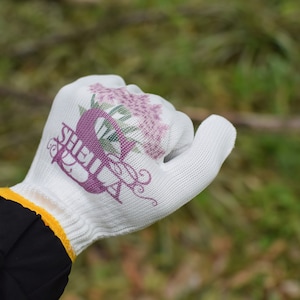 May include: White gardening glove with a floral design and the name "SHEILA" in purple script. The glove has a yellow trim and is set against a blurred green background.