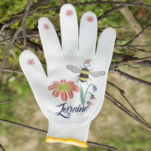 May include: White gardening glove with a yellow trim, featuring a bee, flower, and leaves design. The glove has the name "Lorraine" printed on it.
