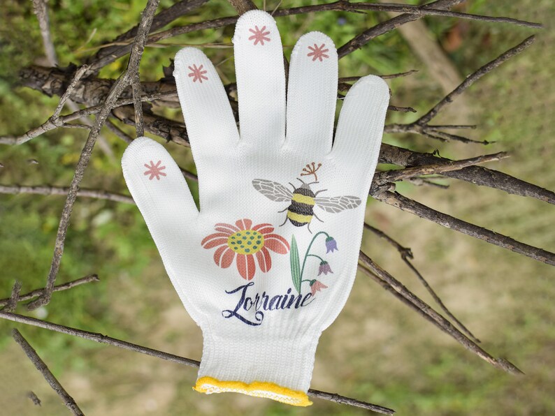 May include: White gardening glove with a yellow trim, featuring a bee, flower, and leaves design. The glove has the name "Lorraine" printed on it.