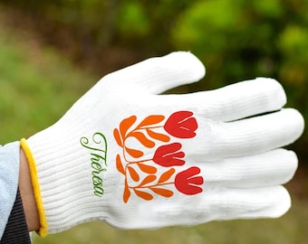 Op de afbeelding: Witte tuinhandschoen met een bloemmotief in rood en oranje, en de naam "Theresa" in groene letters. De handschoen heeft een gele rand.