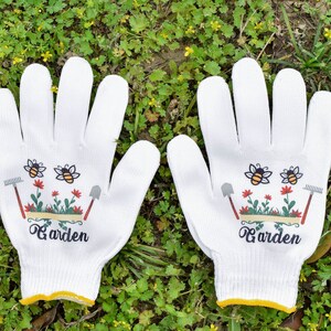 Puede incluir: Un par de guantes de jardinería blancos con ribete amarillo. Los guantes tienen un diseño de abejas y flores y el texto "Garden" impreso en ellos.