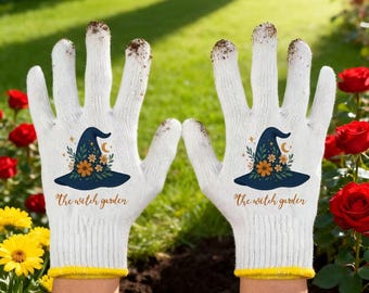 Op de afbeelding: Witte tuinhandschoenen met een gele rand. Elke handschoen heeft een marineblauw heksenhoedontwerp met oranje bloemen en de tekst "the witch garden". De handschoenen zijn bedekt met aarde.