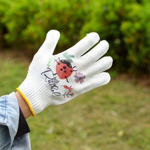 K&ouml;nnte beinhalten: Wei&szlig;e Gartenhandschuhe mit einem Marienk&auml;fer- und Blumendesign. Die Handschuhe haben den Schriftzug "Ditsy" aufgedruckt.