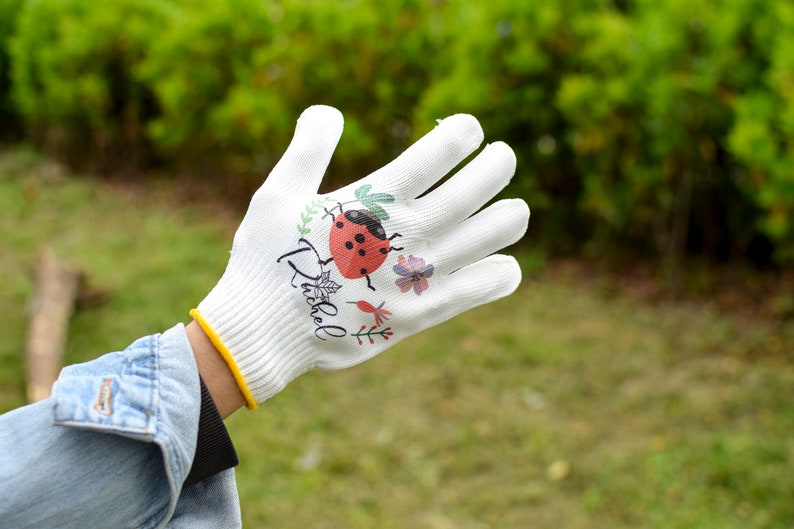 K&ouml;nnte beinhalten: Wei&szlig;e Gartenhandschuhe mit einem Marienk&auml;fer- und Blumendesign. Die Handschuhe haben den Schriftzug "Ditsy" aufgedruckt.