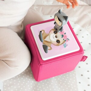 May include: A pink fabric cube with a sloth and a raccoon character on the top. The cube has a white background with pink flowers and butterflies. A baby is playing with the cube.
