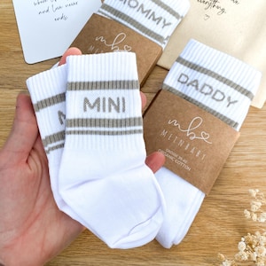 May include: Three pairs of white crew socks with grey stripes and text. One pair features the word MOMMY, one pair features the word DADDY, and one pair features the word MINI. Each pair is wrapped in a brown paper band with the text MEINBABY.