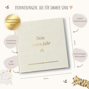 Op de afbeelding: Een cr&egrave;mekleurig hardcover boek met gouden letters die "Dein erstes Jahr" (Je eerste jaar) en een regenboog symbool lezen. Het boek heeft 136 pagina's en meet 21 cm bij 21 cm. Het heeft een gouden reli&euml;f ontwerp en een stevige binding.