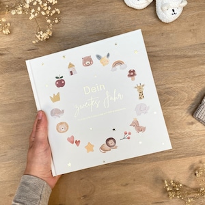 Könnte beinhalten: Ein weißes Buch mit einem goldenen Titel, der "Dein zweites Jahr" lautet. Das Buch ist mit goldenen Sternen und verschiedenen Illustrationen von Tieren und Objekten verziert, darunter eine Krone, ein Wal, ein Löwe, eine Giraffe, ein Pilz, ein Regenbogen, ein Haus, ein roter Apfel und ein Dackel.