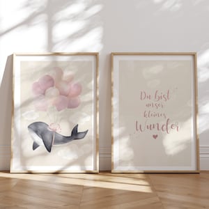 Op de afbeelding: Twee ingelijste prints met een aquarel-illustratie van een walvis die roze ballonnen vasthoudt en de tekst "Du bist unser kleines Wunder" in roze letters.