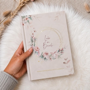 May include: A light beige hardcover book featuring a floral wreath illustration in shades of pink and green, with gold foil accents. The text "Liebe im Bauch" is printed in the center, and "MEIN SCHWANGERSCHAFTSTAGEBUCH" is printed at the bottom.