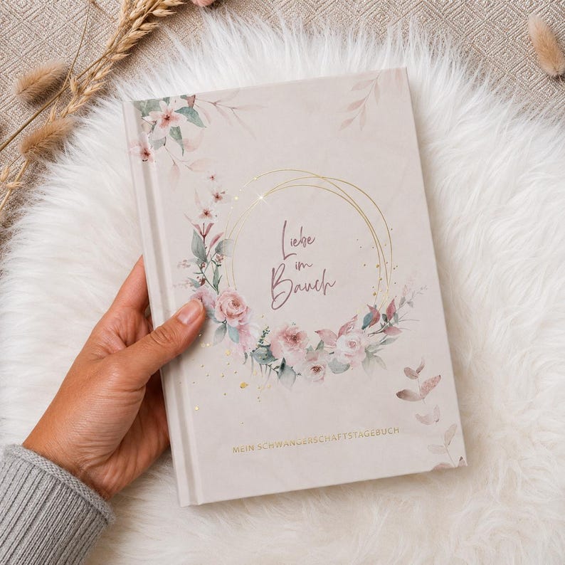 May include: A light beige hardcover book featuring a floral wreath illustration in shades of pink and green, with gold foil accents. The text "Liebe im Bauch" is printed in the center, and "MEIN SCHWANGERSCHAFTSTAGEBUCH" is printed at the bottom.